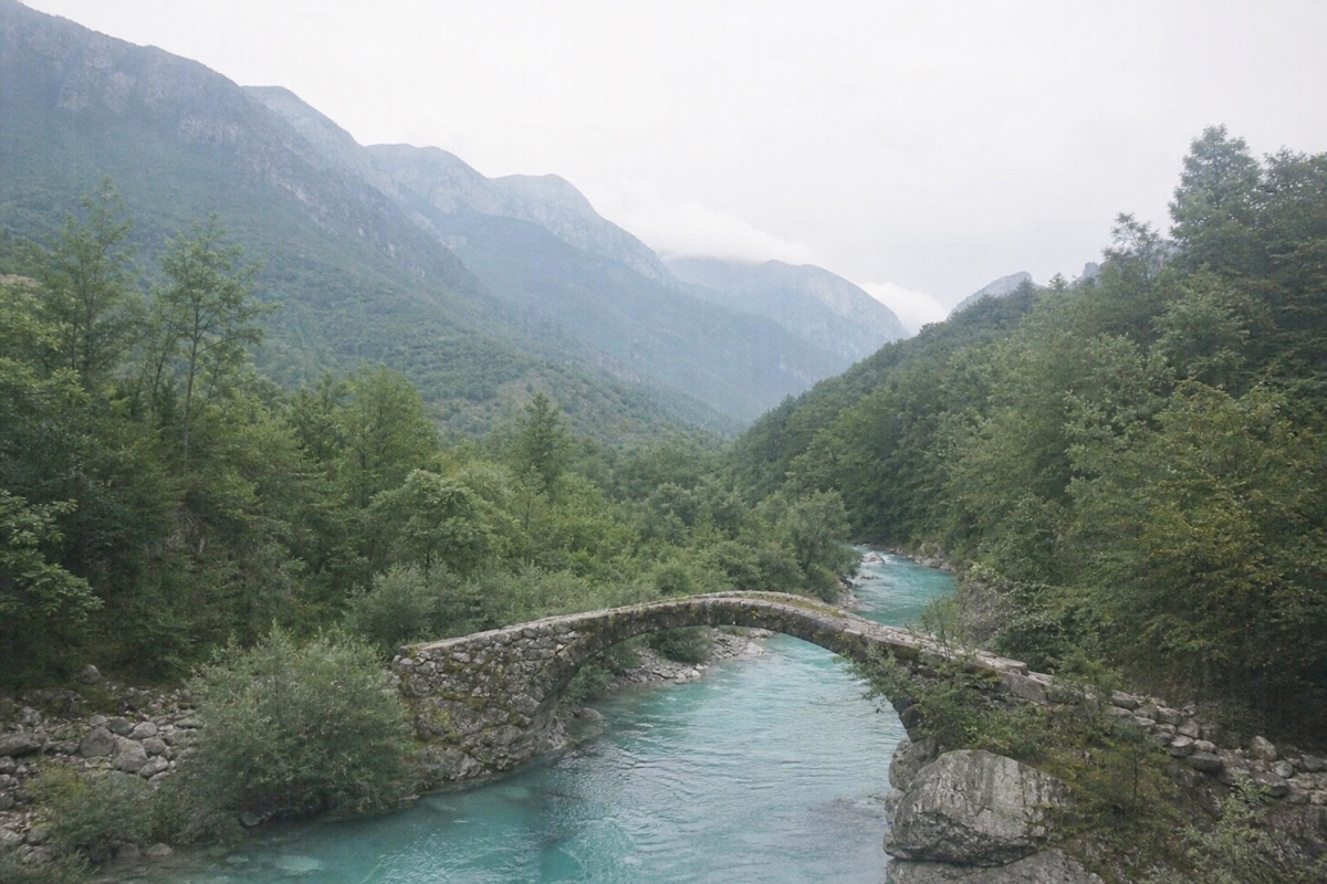 A bright mountain valley and stone bridge veiled by haze, flat colour and soft detail