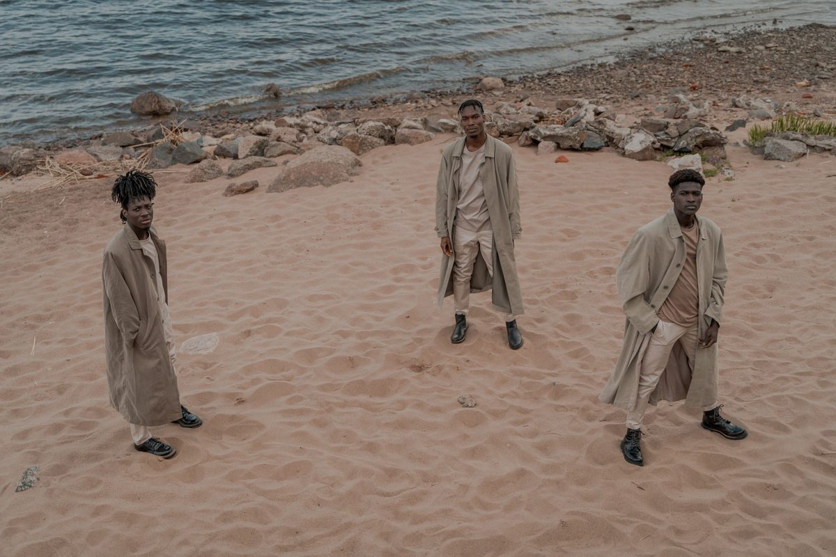 Three men in matching tan trench coats spaced across a sandy shore, the third of them standing on the right
