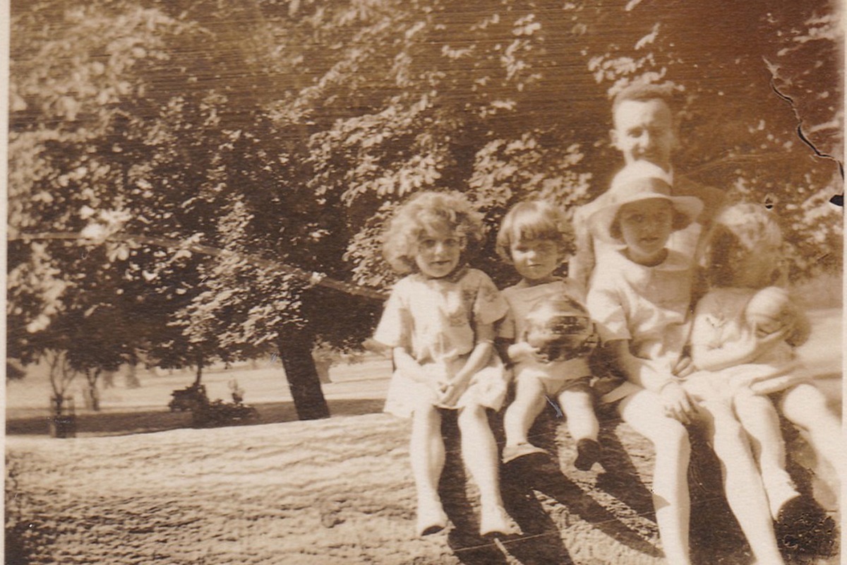 A faded sepia print of children on a bench with an adult behind them, torn at one corner and stained down one edge